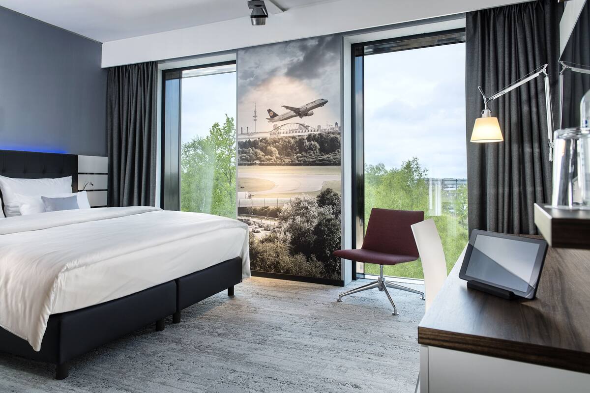 Een moderne hotelkamer met een groot bed, een bureau met een laptop en een uitzicht op een stadsbeeld door grote ramen.