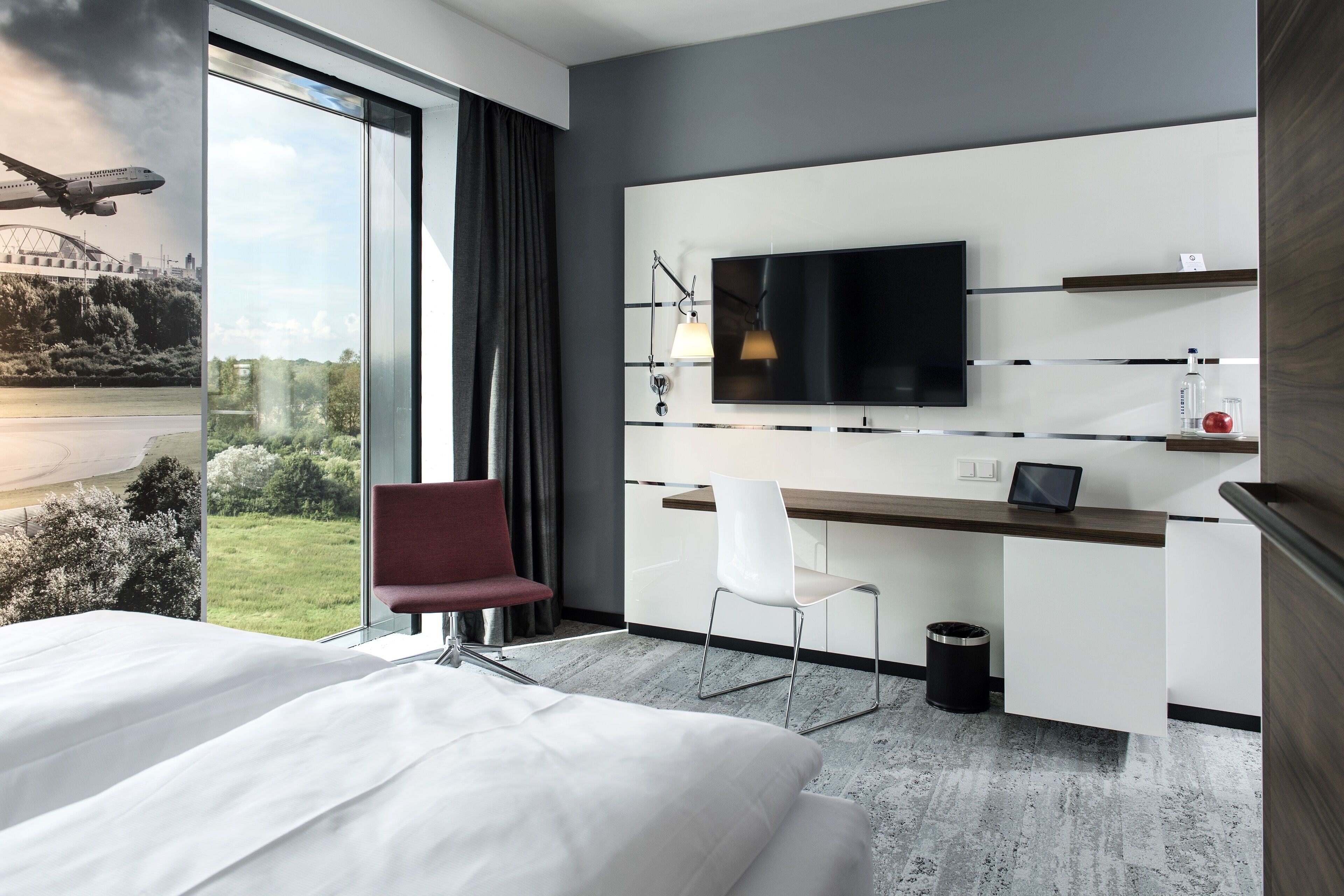 A modern hotel room with a large bed, a desk with a chair, a television, and a view of a bridge and greenery.