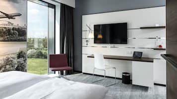 A modern hotel room with a large bed, a desk with a chair, a television, and a view of a bridge and greenery.