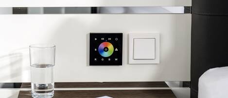 A modern hotel room with a bedside table, a glass of water, a pen, a notepad, a color-changing light control panel, and a shower head.