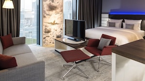 A modern hotel room with a large bed, a desk with a flat-screen TV, a sofa, and a chair.