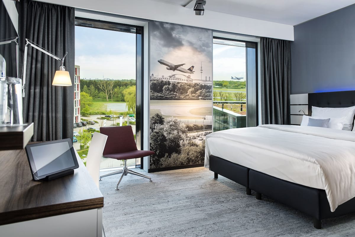 Een moderne hotelkamer met een groot bed, een bureau met een laptop, een stoel en uitzicht op een brug en een vliegveld.