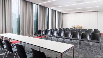 A conference room with a long table and rows of chairs, large windows with curtains, and a carpeted floor.