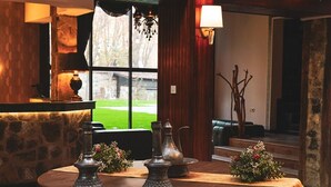 Ein runder Tisch mit drei Metallkerzenhaltern und einer Blumenvase, aufgestellt auf einem Holztisch in einem Raum mit Steinwänden, einem Fenster und einer Kristallleuchter.