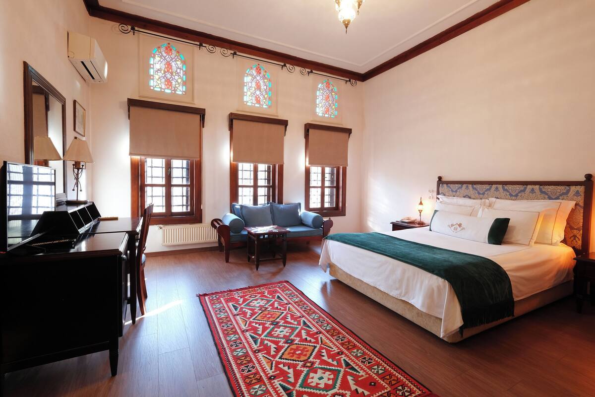 A hotel room with a piano, a sofa, a bed with a green blanket, and a red patterned rug.
