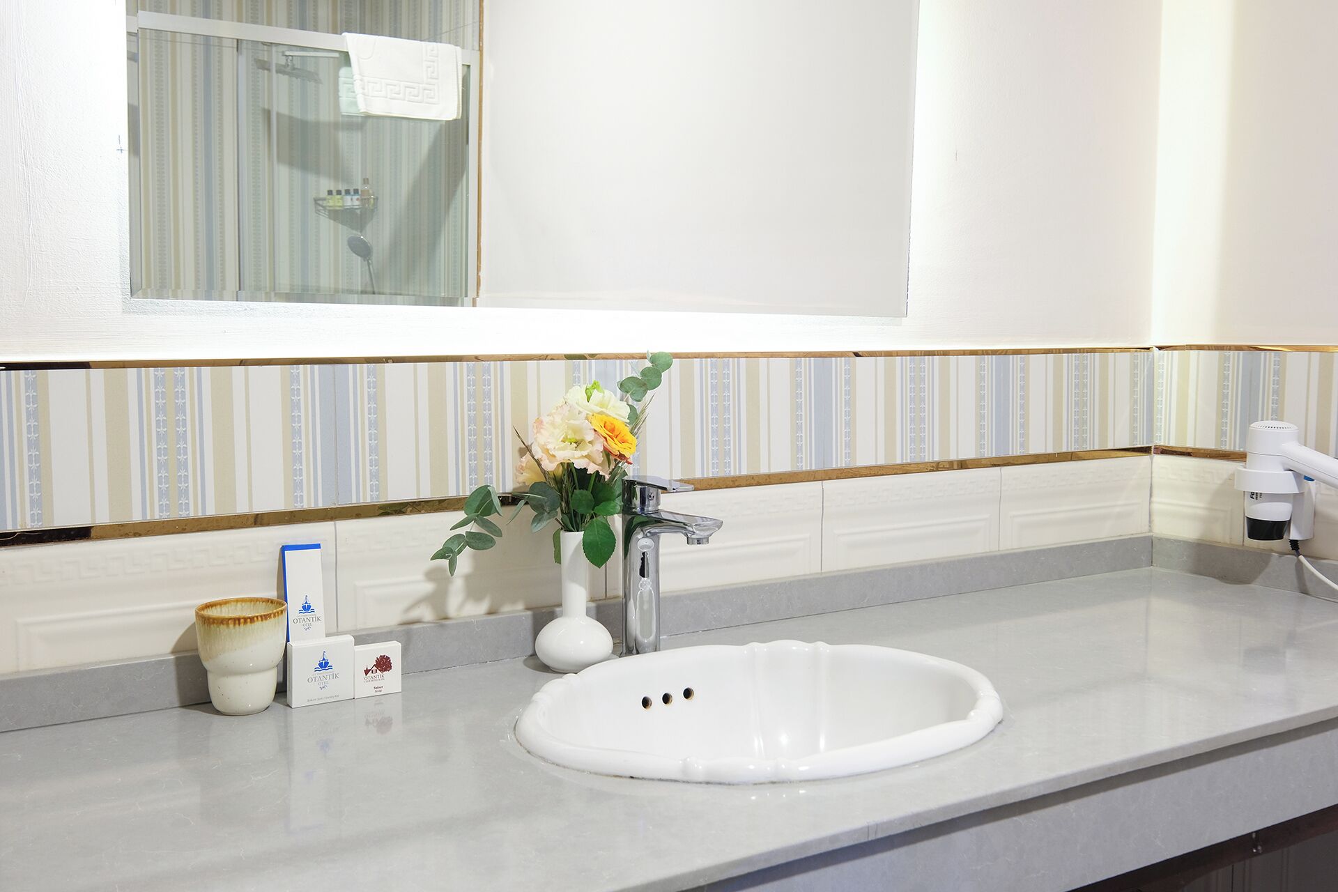 Casa de banho com lavatório branco, espelho e um vaso com flores. Há artigos de higiene pessoal no balcão.