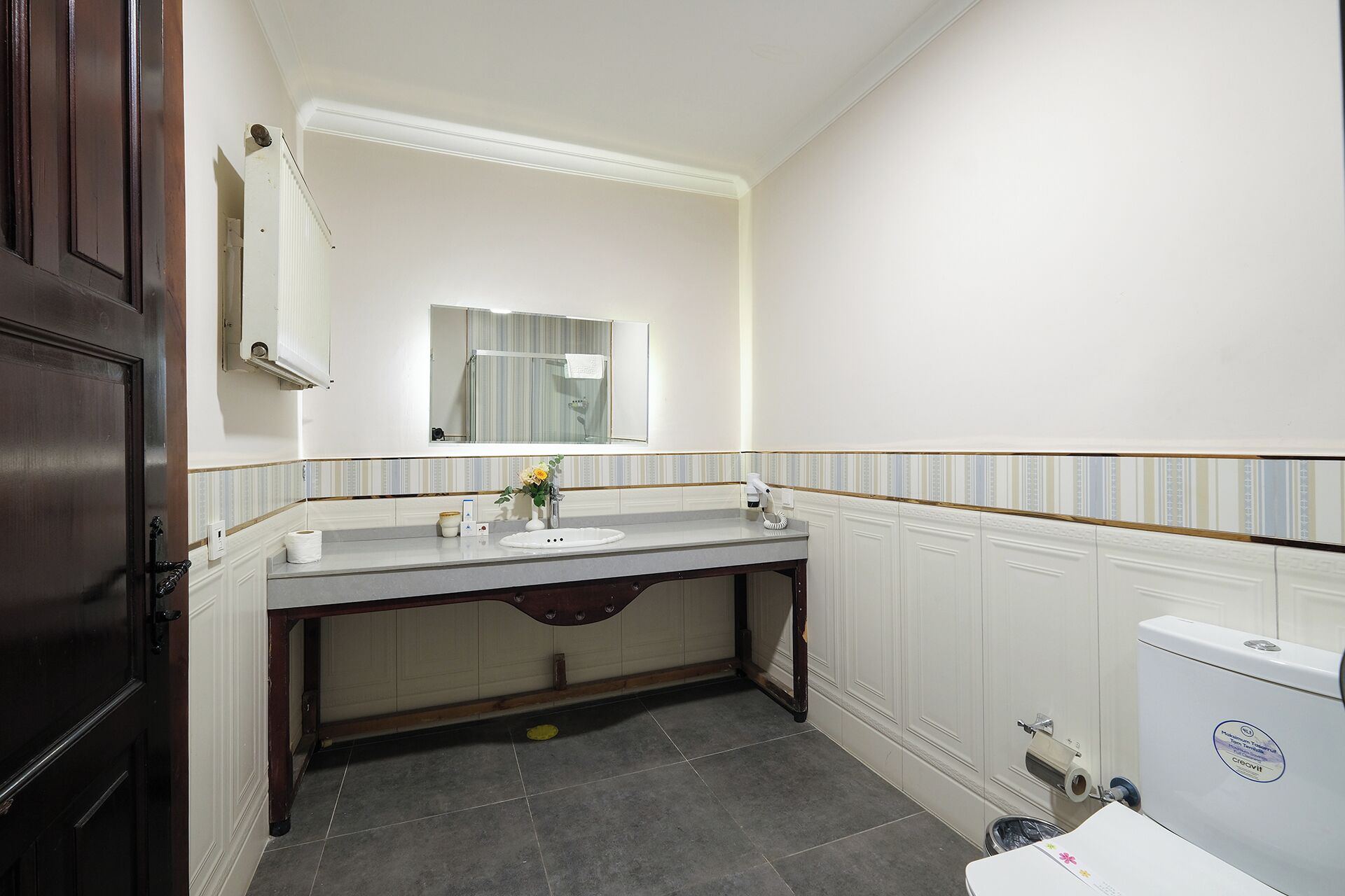Un baño con puerta de madera, un lavabo blanco con un espejo encima, una toalla colgada en la pared y un inodoro.