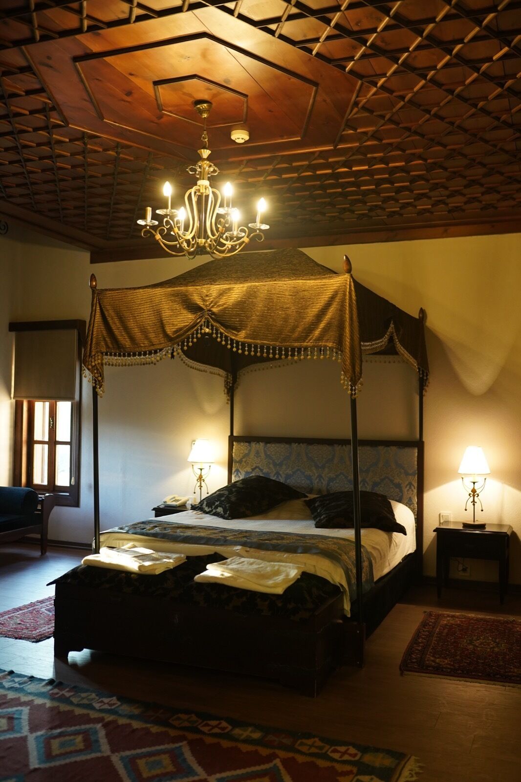 A four-poster bed with a canopy, a chandelier, and a bedside table with a lamp.