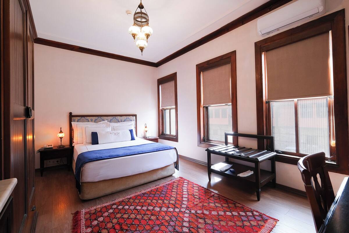 A hotel room with a bed, a desk, a chair, a red patterned rug, and a chandelier.