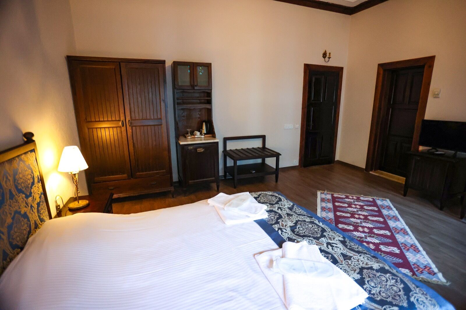 A hotel room with a large bed, a wooden wardrobe, a small table with a lamp, a television, and a patterned rug.