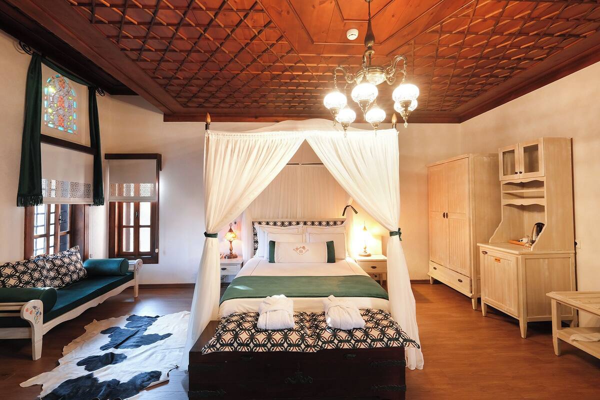 A room with a four-poster bed, a wooden ceiling, a chandelier, a wooden wardrobe, and a window with a decorative view.
