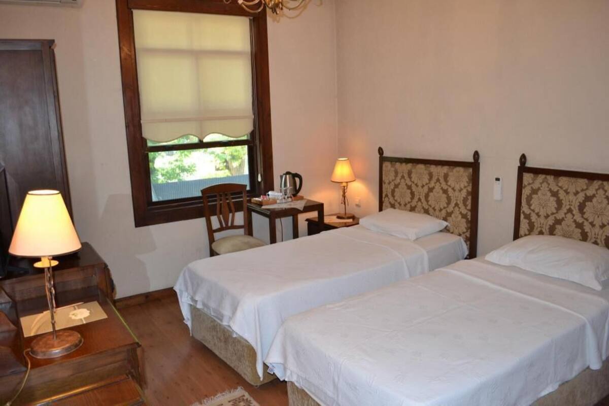 A hotel room with two beds, a desk with a lamp, a chair, a window with blinds, and a small table with a kettle.