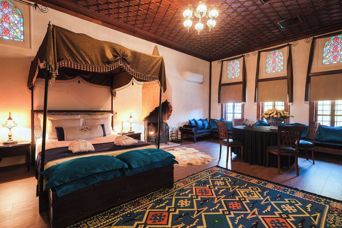 A four-poster bed with a canopy, a fireplace, a dining area with a table and chairs, and a patterned rug.