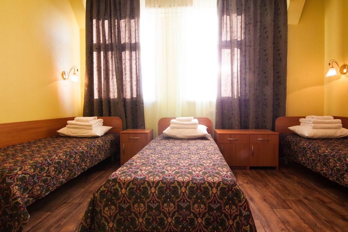 Habitación de hotel con tres camas individuales, cada una con una colcha estampada y una toalla doblada. La habitación tiene suelo de madera, una ventana con cortinas de encaje y luces fijadas a la pared.