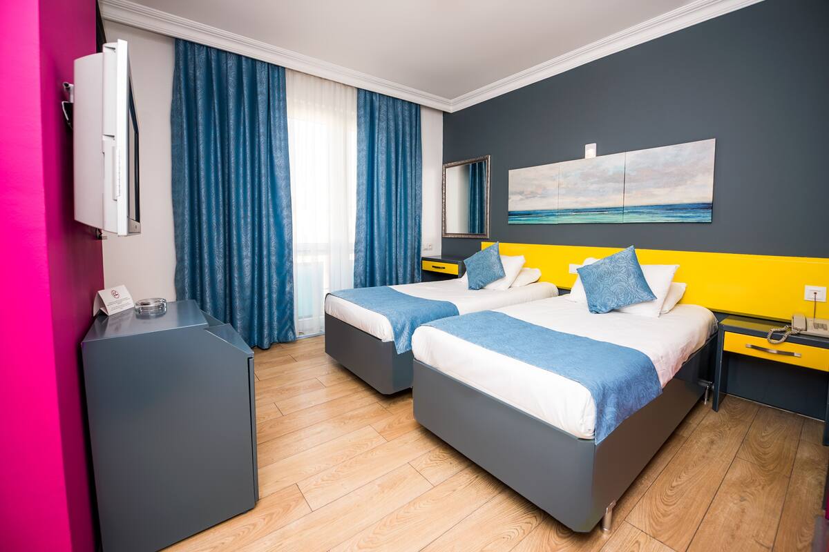 A hotel room with two beds, a desk, a mirror, and a painting of a beach.