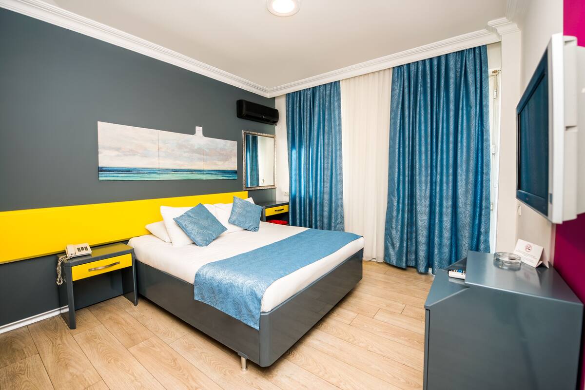 A modern hotel room with a large bed, a bedside table with a phone, a desk with a computer, and a large window with curtains.