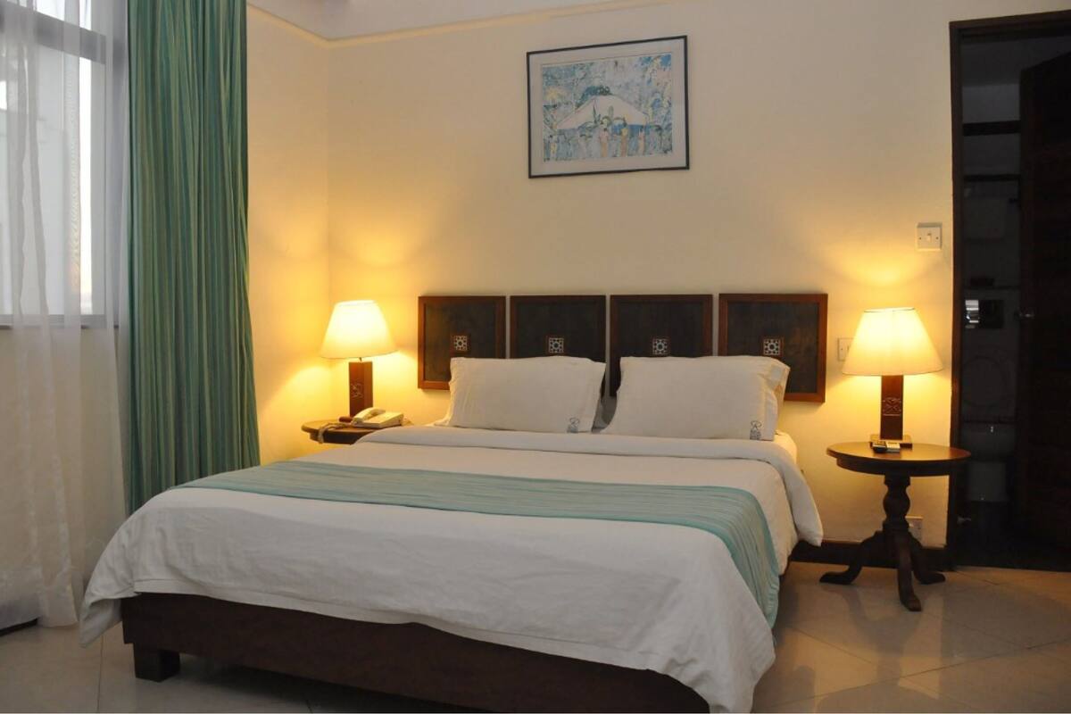 A hotel room with a bed, two bedside lamps, a nightstand, a telephone, a painting on the wall, and a green curtain.