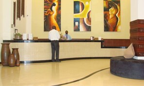 A reception desk with a receptionist, a large abstract painting, and a reception area with a tiled floor and a seating area.