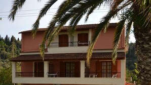 A two-story building with a balcony on the upper floor, a small garden area with a fountain, and a paved entrance. - Anna Apartments (Corfu)