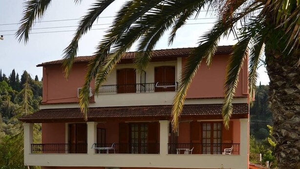A two-story building with a balcony on the upper floor, a small garden area with a fountain, and a paved entrance. - Anna Apartments (Corfu)