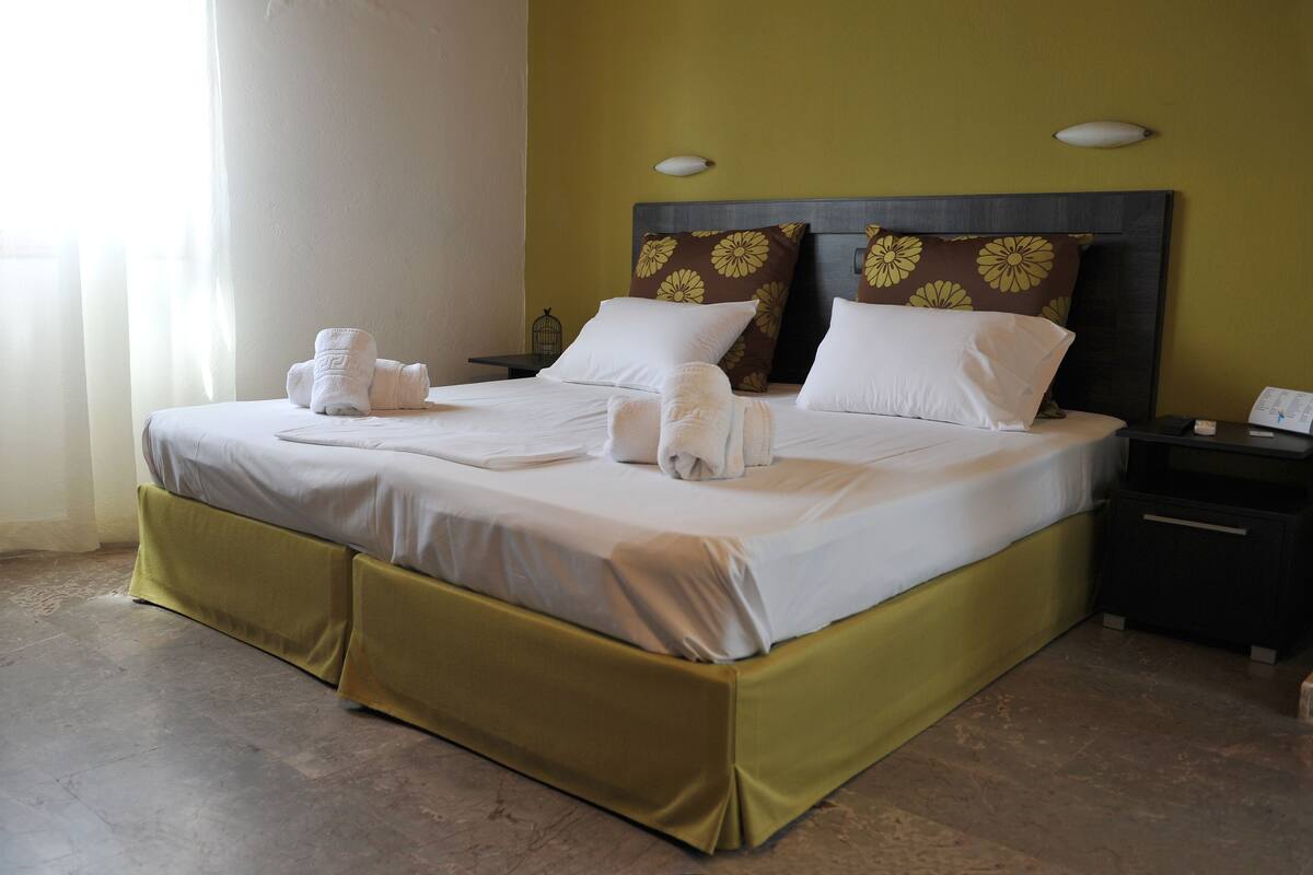 A neatly made bed with white linens and towels, a dark headboard with floral pillows, and a nightstand with a lamp and a phone.