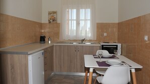 A compact kitchen with a refrigerator, microwave, and sink. There is a window with curtains, a small table with chairs, and a coffee maker. - Anna Apartments (Corfu)