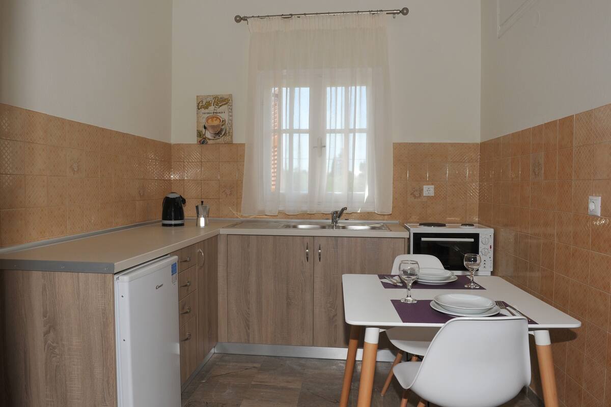 A compact kitchen with a refrigerator, microwave, and sink. There is a window with curtains, a small table with chairs, and a coffee maker.