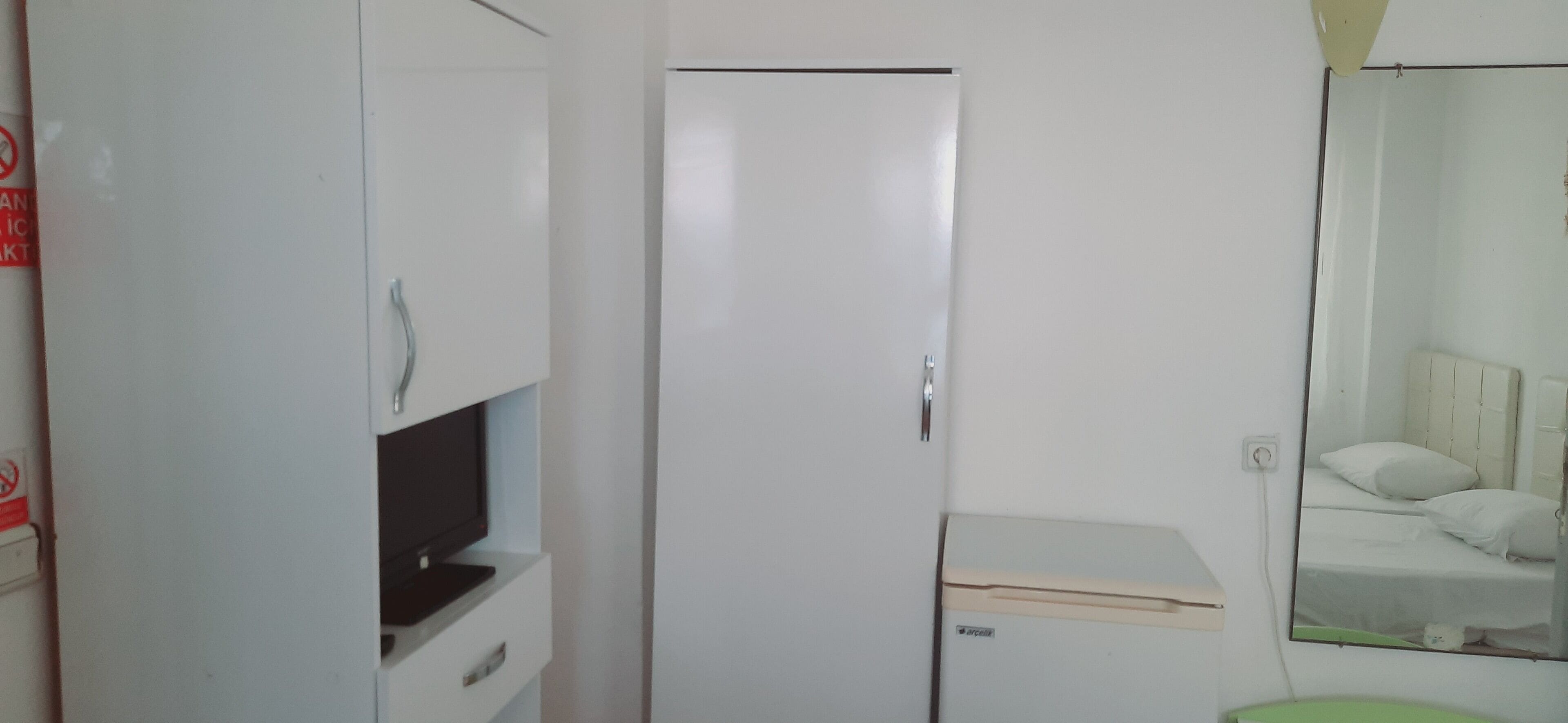 Een kleine, compacte kamer met een witte kast, een gesloten deur en een spiegel die een bed weerspiegelt.