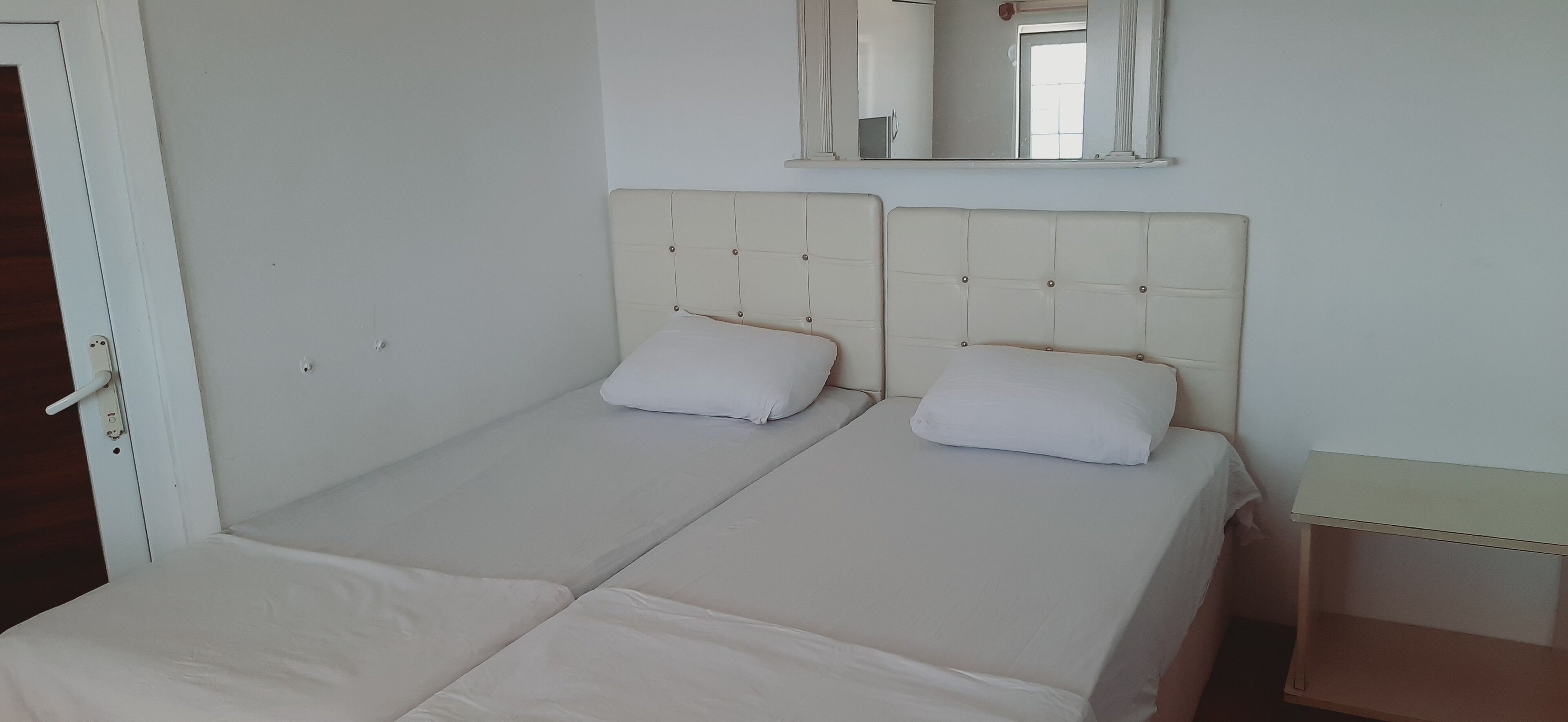 A room with two single beds, a small bedside table, a mirror above the beds, and a door on the left.