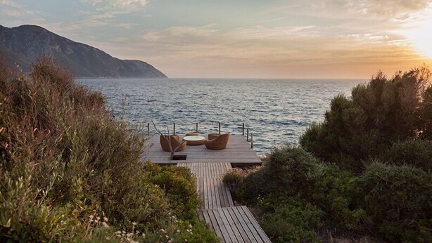 Une terrasse en bois avec des sièges extérieurs, donnant sur une étendue d’eau et sur une montagne au loin.