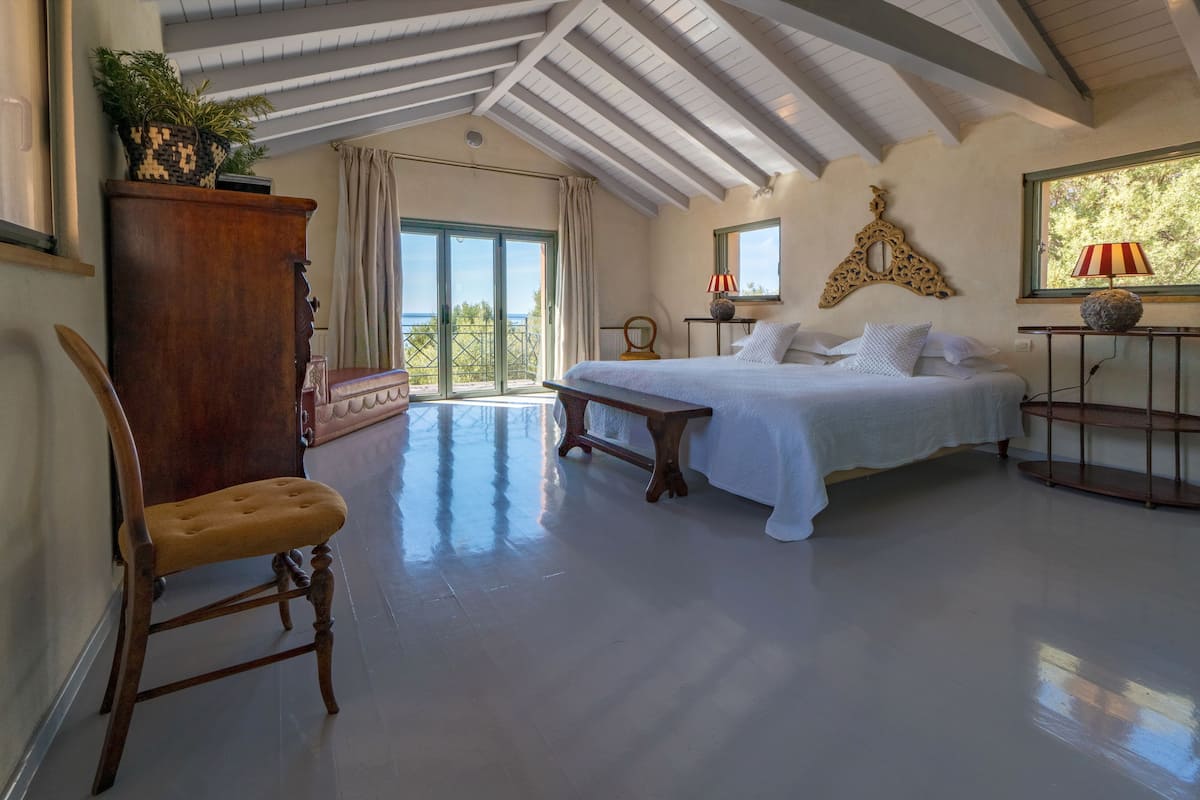 A spacious bedroom with a large bed, a wooden chair, a nightstand, and a view of the outdoors through a sliding door.