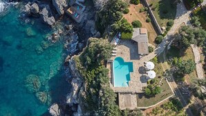 Vue aérienne d’une propriété située sur la côte, avec une piscine, un terrain rocheux et une végétation luxuriante.