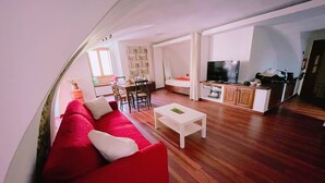 A living room with a red sofa, white coffee table, and a TV. There is a dining area with a table and chairs, and a kitchen area with cabinets and a TV. - Chiostro San Marco (Tarquinia)