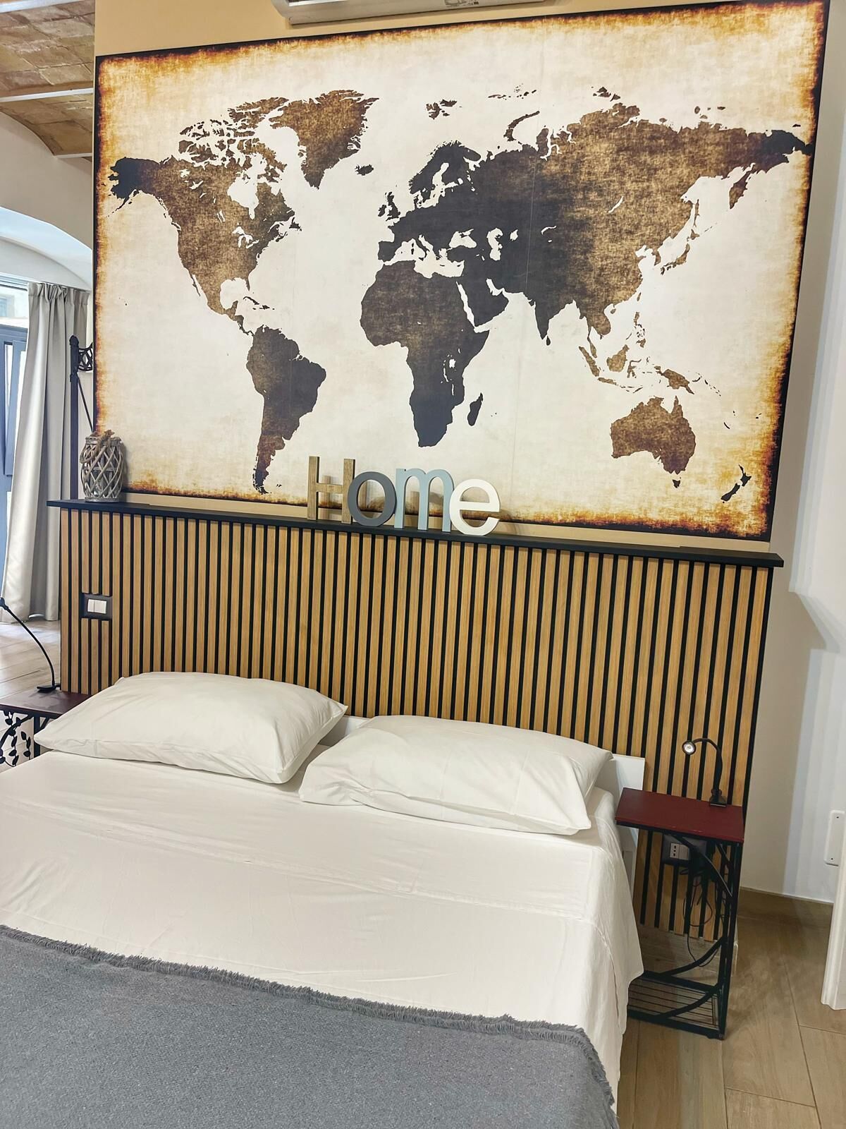 A bed with a world map above it, a wooden slat headboard, and a small table with a lamp.