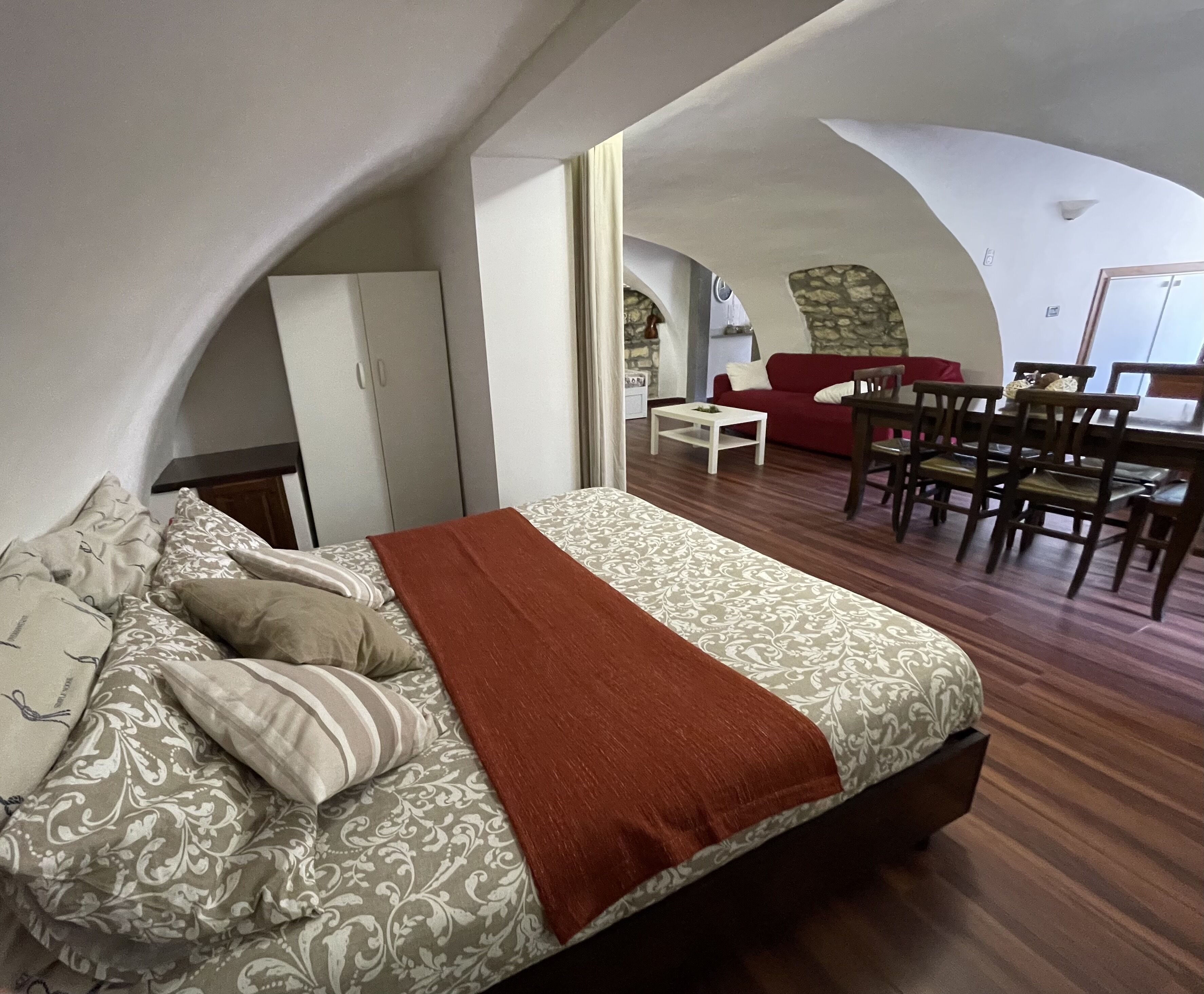 Une chambre avec un grand lit, une couverture rouge et une couette à motifs. On y trouve également un coin repas avec un canapé rouge et des chaises en bois.