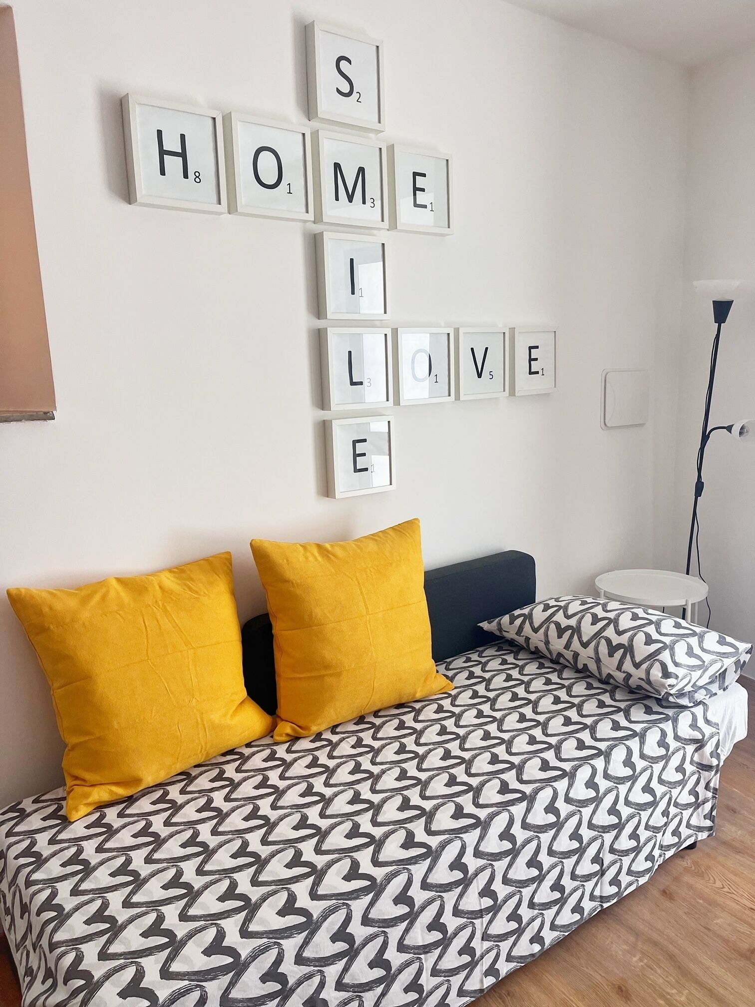 Una cama con cubrecama estampado, dos almohadas, una mesita de noche y una pared con fichas de Scrabble que deletrean 'HOME LOVE'.