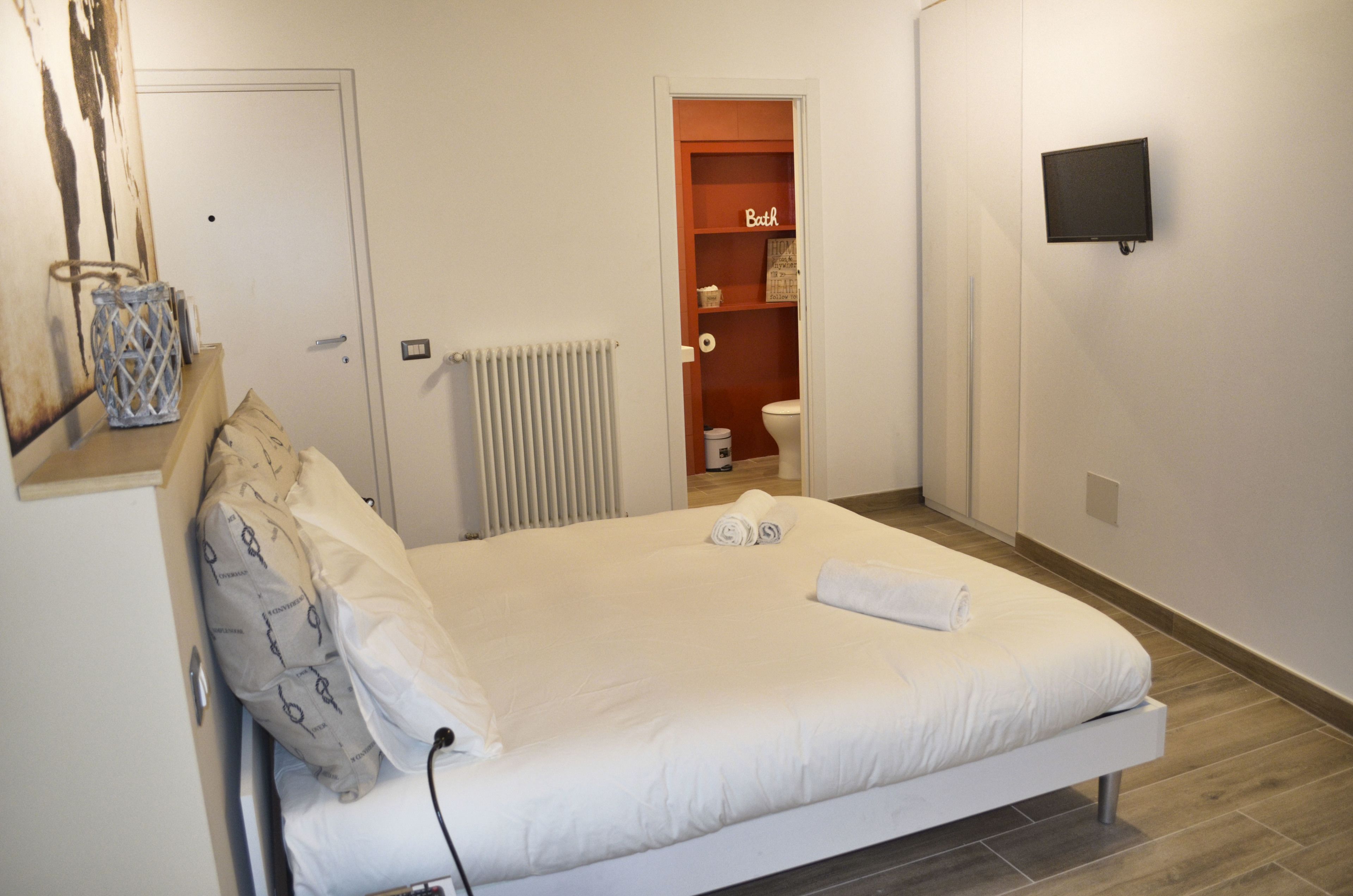 A neatly made bed with white linens and a rolled towel, a white radiator, a television mounted on the wall, and a small shelf with a sign reading "Bath".