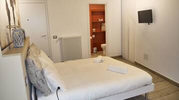 A neatly made bed with white linens and a rolled towel, a white radiator, a television mounted on the wall, and a small shelf with a sign reading "Bath".