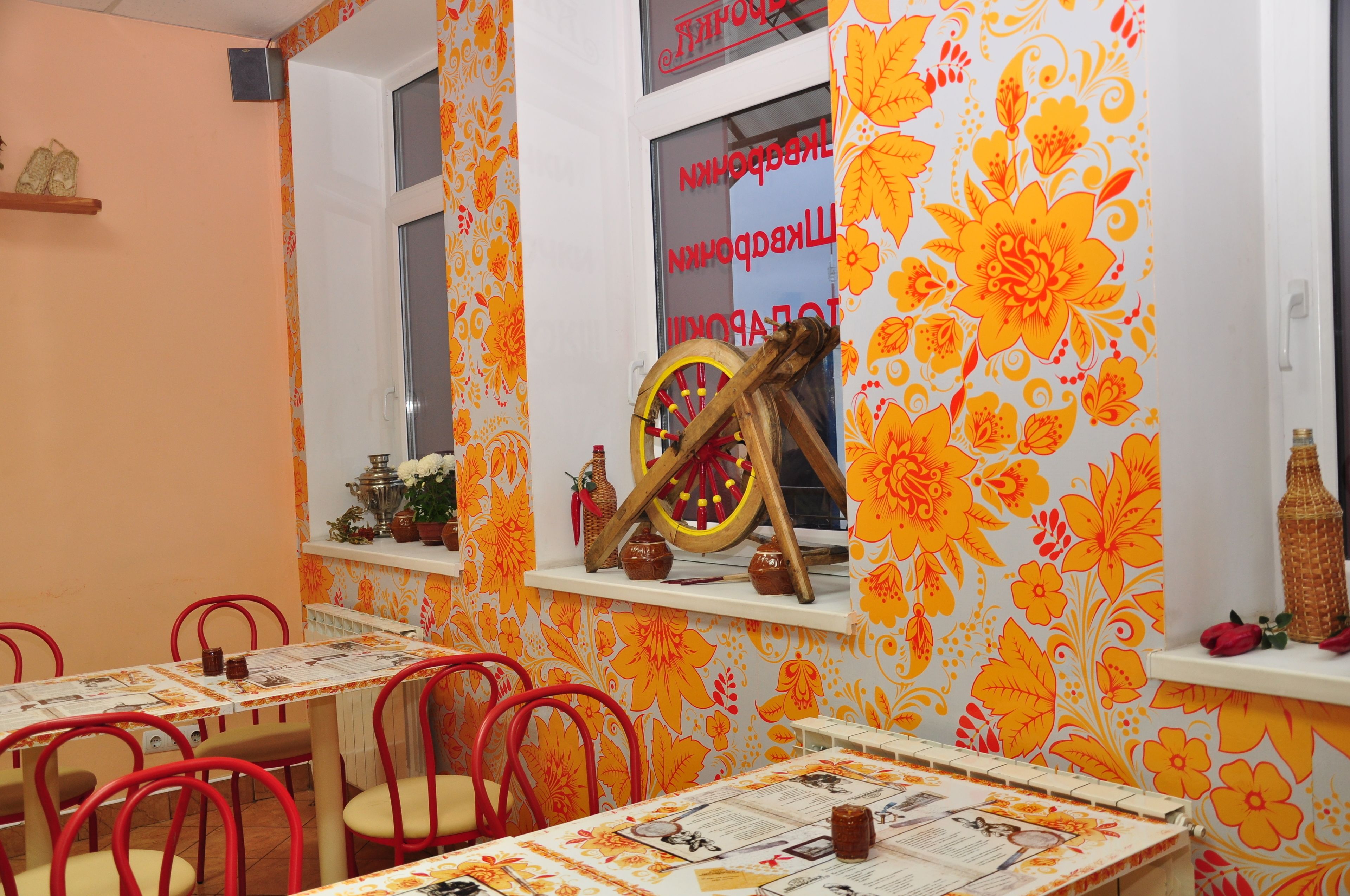 A dining area with tables and chairs, a window with a floral pattern, and a wooden wheel decoration.