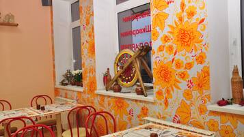 A dining area with tables and chairs, a window with a floral pattern, and a wooden wheel decoration.