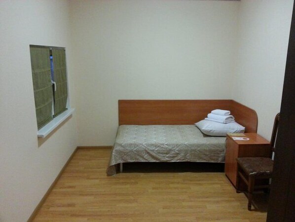 Una habitación pequeña con cama individual, cabecera y mesita de noche de madera, y una silla. Hay una ventana con cortina recogida a un lado.