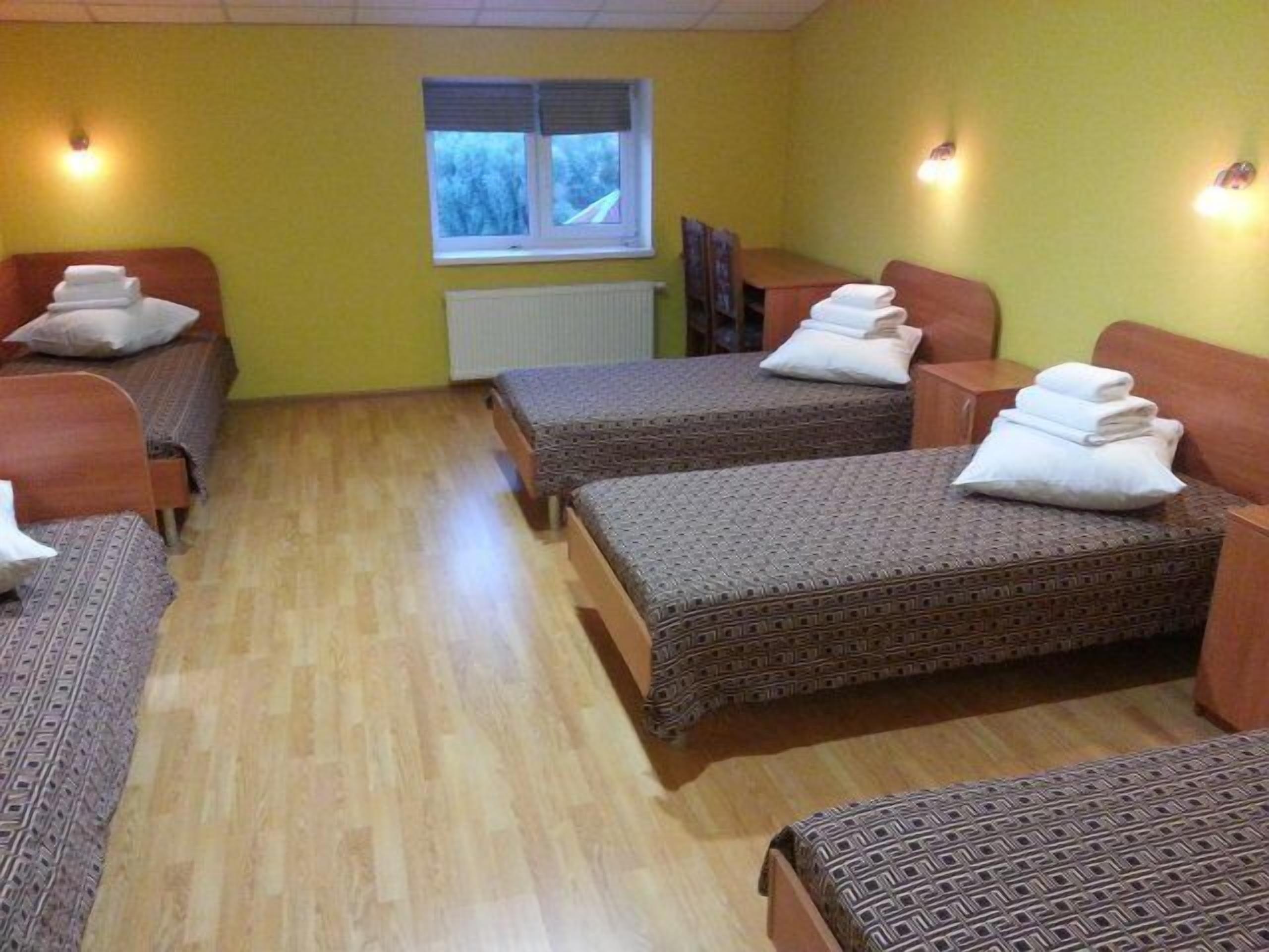 A room with two single beds, each with a patterned bedspread and a folded towel. There is a window with blinds, a radiator, and a wooden table with a chair.