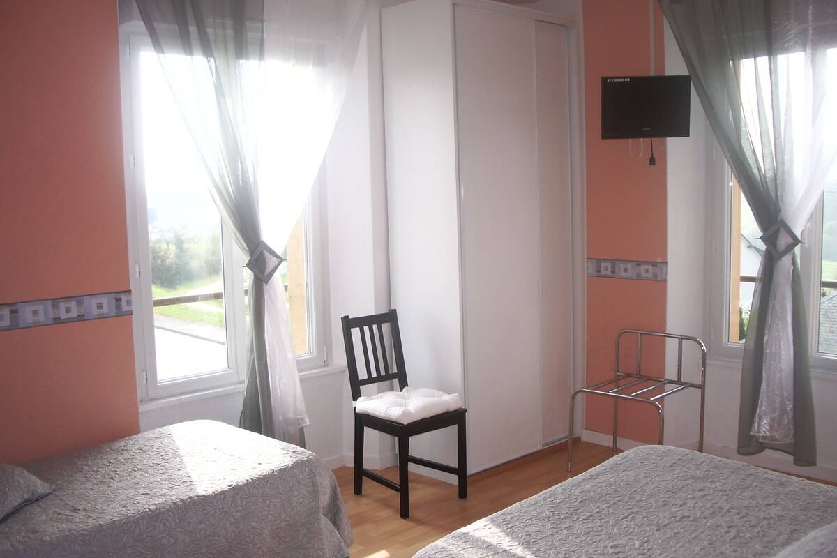 Una habitación con dos camas, una silla, un televisor y un ventanal con cortinas.
