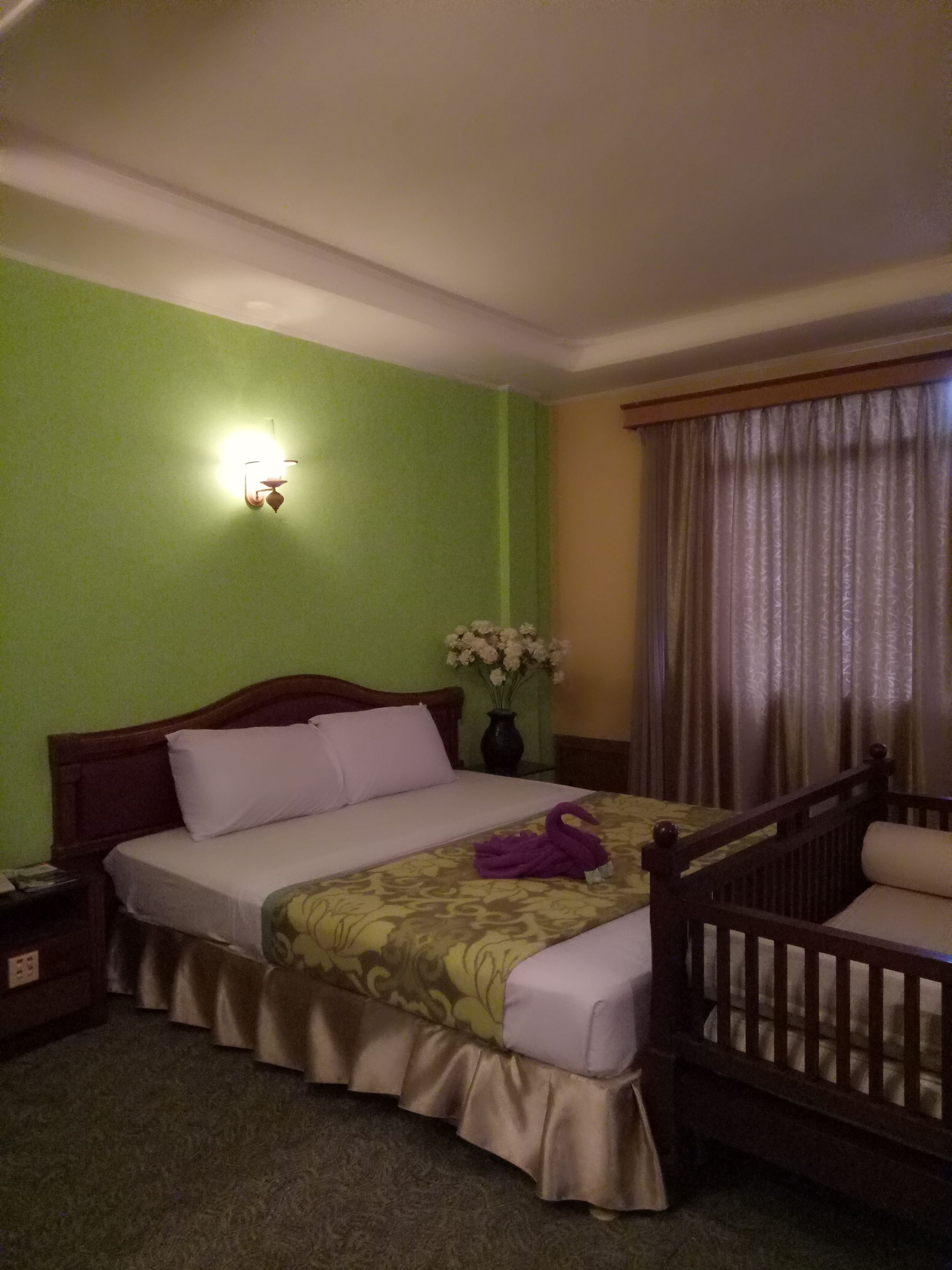 Una habitación de hotel con cama, mesita de noche, un jarrón con flores y una ventana con cortinas.