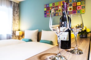 Ein Hotelzimmer mit einem Bett, einem Holztisch mit einer Weinflasche und zwei Gläsern sowie einem farbenfrohen Wandbild.