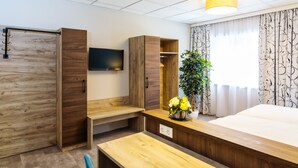 Hotellihuone, jossa on sänky, seinälle asennettu televisio, puinen työpöytä, vaatekaappi ja ruukussa oleva kasvi.