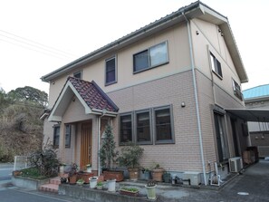 Une maison à deux étages avec une façade en briques, un toit en tuiles et de nombreuses fenêtres. On peut voir des plantes en pot sur le porche et une zone abritée sur le côté droit.