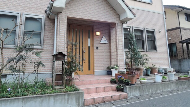Een gebouw met twee verdiepingen, een beige gevel, een houten deur en een klein portaal met plantenpotten.