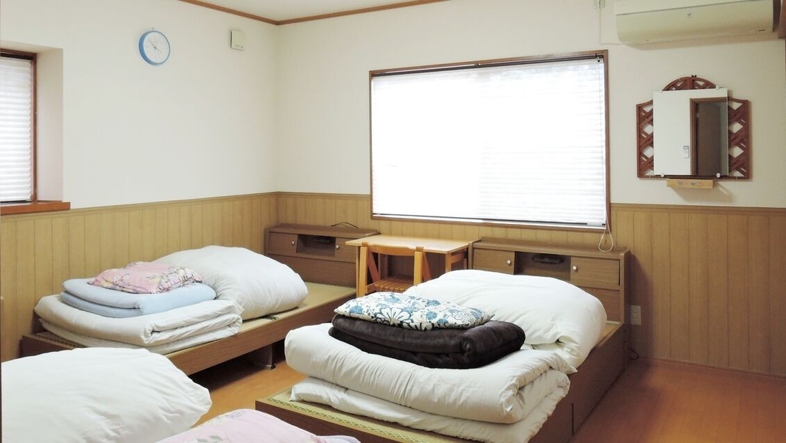 A room with two beds, a small table, a window with blinds, a clock, and a mirror on the wall.