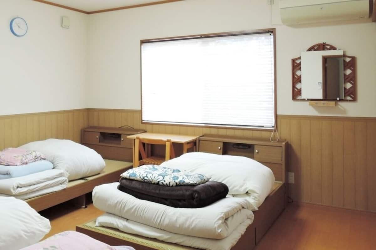 Ein Zimmer mit zwei Betten, einem kleinen Tisch, einem Fenster mit Jalousien, einer Uhr und einem Spiegel an der Wand.